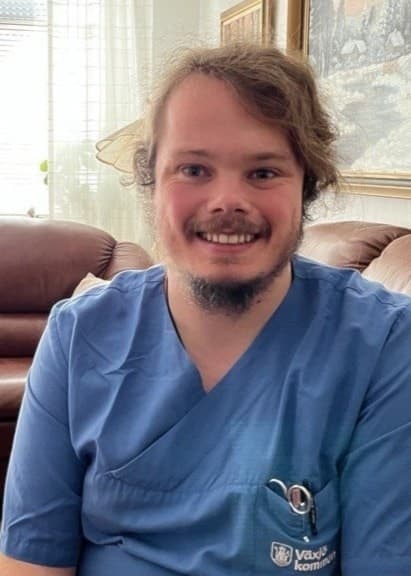 Ted Fransén sitter i en brun skinnsoffa i ett vardagsrum med ljusinsläpp från ett fönster. Han bär blå sjukvårdskläder och omgivningen inkluderar frukt på ett soffbord.