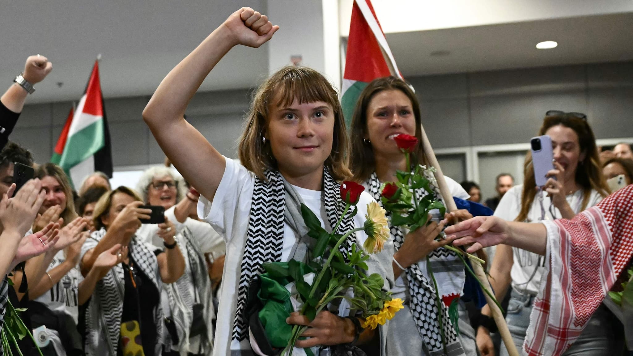 Greta Thunberg håller handen över hjärtat och är klädd i en vit tröja med en svartvit palestinsk halsduk, omgiven av personer vid ankomst till Atens internationella flygplats efter deportation från Israel.