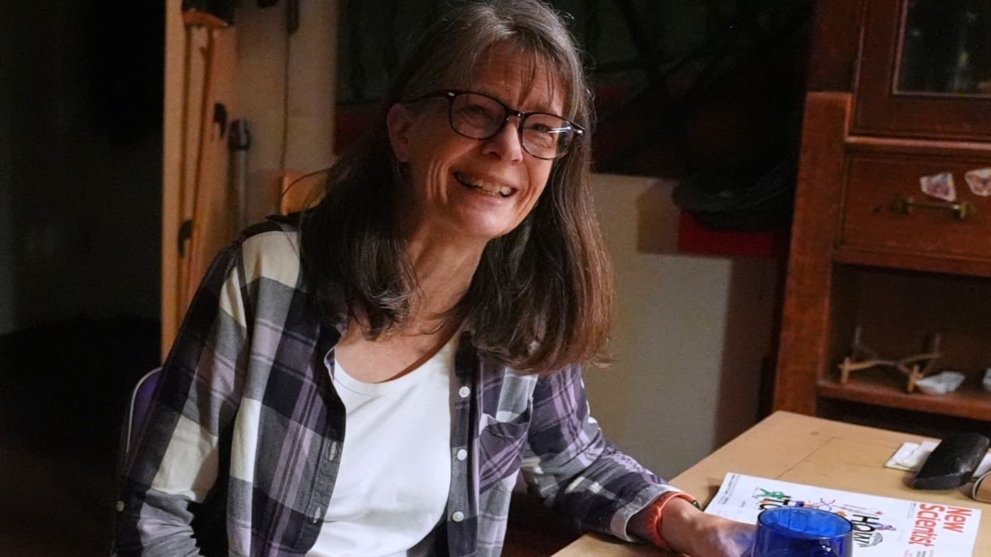 Mary E. Brunkow, klädd i en rutiga skjorta, sitter vid ett köksbord med en laptop, mobil och kaffemugg, och ler efter att ha fått veta att hon vunnit Nobelpriset i medicin för forskning inom immunologi.